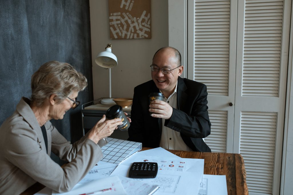 A imagem retrata uma mulher mais velha, branca, e um homem mais velho, asiático, tomando café em uma sala de reuniões.
